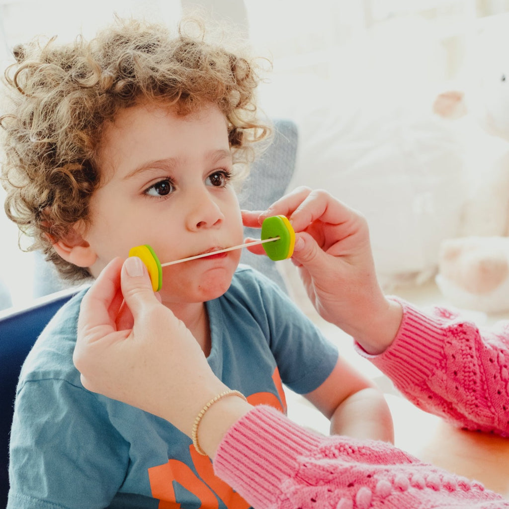 A child with curly hair uses Lip Gym™ while an adult demonstrates the exercise with green tools.