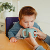 A child drinking from a light blue TalkTools Itsy Cup with handles, held by an adult, seated at a wooden table.